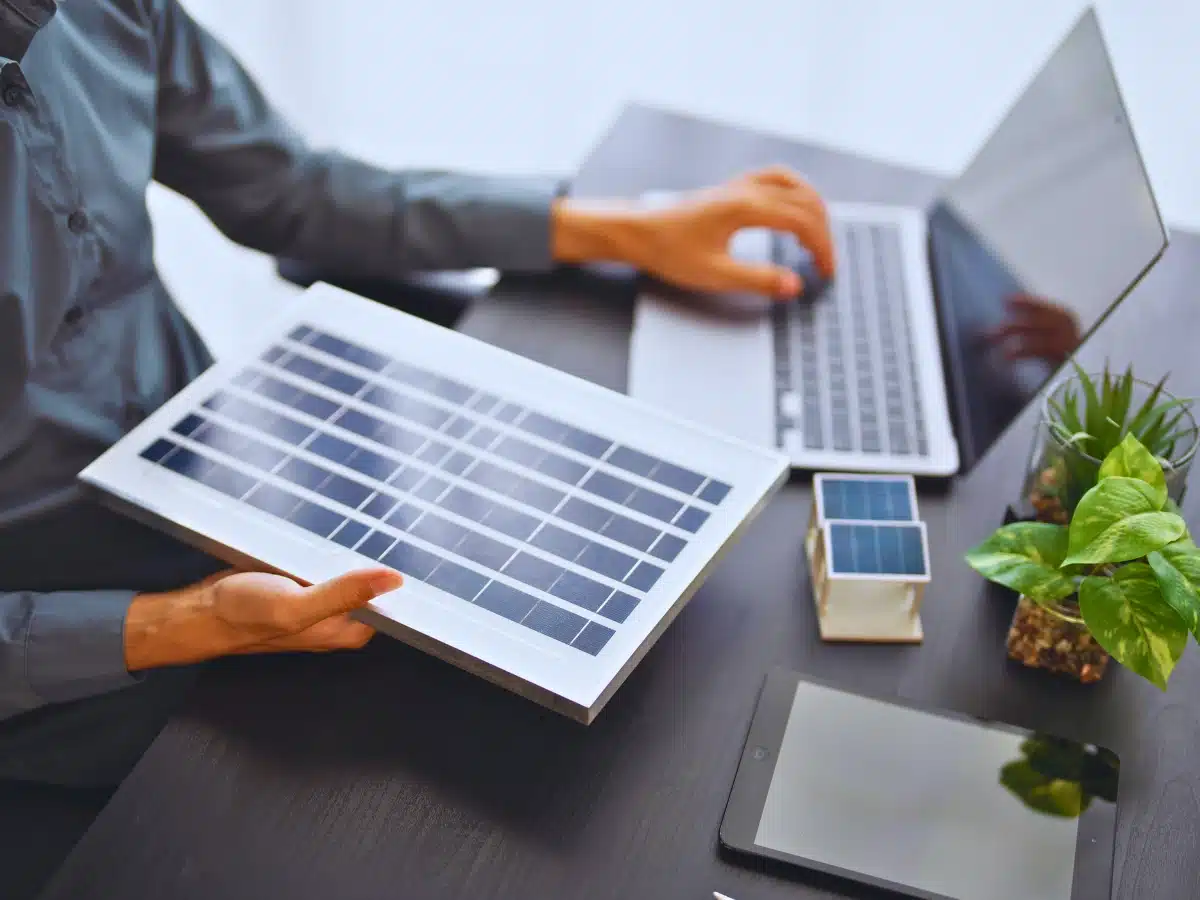 Engineer holding a small solar panel prototype while working on a laptop at a desk